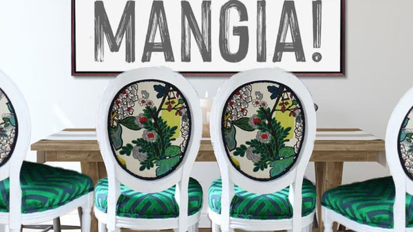 2 white round back dining chairs with a green, white and black, Asian inspired print.  The seat of the chair has a green and blue velvet geometric patterned fabric.  The chairs are up against a wooden table with a sign behind it that says “Mangia!”
